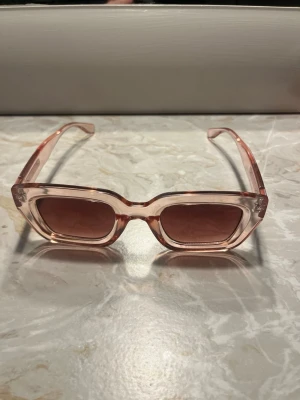 Rosa transparenta solglasögon - Snygga solglasögon med transparent rosa bågar och fyrkantig form. Glasen är lätt tonade och bågarna har en modern, chunky design. Perfekta för dig som vill sticka ut med en trendig accessoar.