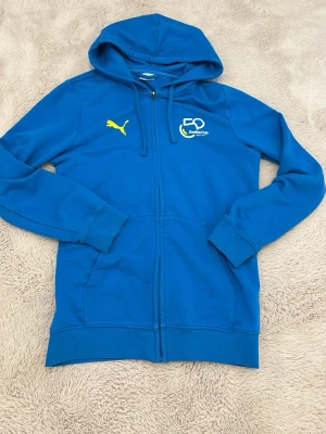 Blå zip-hoodie från Puma Gothia Cup - Blå hoodie med dragkedja från Puma, Gothia Cup edition. Har gul Puma-logga och Gothia Cup 50-års tryck på bröstet. Utrustad med huva och två fickor framtill. Perfekt för dig som gillar sportig stil. 