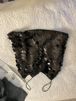 Svart paljett tubtopp - Säljer en svart tubtopp täckt med stora glittriga paljetter. Toppen är axelbandslös och har en croppad passform, perfekt för att sticka ut på festen. Tunn och lätt, med elastisk kant upptill för att sitta på plats.