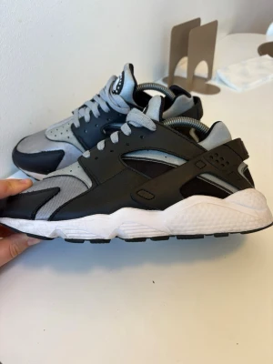 Nike Air Huarache svart och grå - Nike Air Huarache sneakers i svart och grått med vit sula. Skorna har en unik design med blandning av mesh och syntetmaterial, samt en stabil hälkappa med Nike-logga. Perfekt för dig som gillar sportig och modern stil. Skorna är specialdesignade på Nikes hemsida
