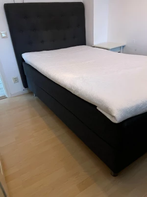 Svart kontinentalsäng med hög gavel - Säljer en stilren svart kontinentalsäng med hög, vadderad huvudgavel och knappar. Sängen har en vit bäddmadrass med diskret mönster och står på svarta ben. Perfekt för dig som vill ha en modern och bekväm säng till sovrummet.