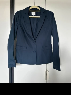 Mörkblå kavaj från Vila i storlek 38 - Snygg mörkblå kavaj från Vila med klassisk krage, två knappar framtill och fickor på sidorna. Figursydd modell som ger en stilren look. Perfekt att matcha med jeans eller kostymbyxor. Materialet känns mjukt och följsamt.