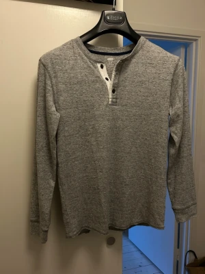 Grå långärmad henley från Old Navy - Säljer en gråmelerad långärmad henleytröja från Old Navy. Tröjan har knappar vid halsen och kontrastfärgad kant i mörkblått. Perfekt för en avslappnad och stilren look. Tillverkad i mjukt bomullsmaterial. Storlek Barn XL-14-16