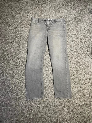 Jack and Jones jeans - Säljer ett par grå raka jeans från Jack and Jones Jeans med klassisk femficksmodell och knappgylf. Jeansen har en ljus tvätt och subtila slitningar för en avslappnad look. Tillverkade i denim med Nudie Jeans logga på bakfickan. W32 L32