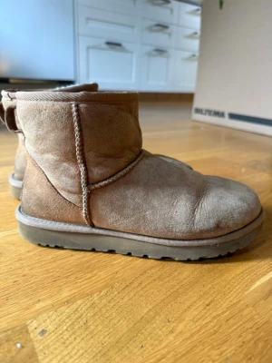 Bruna UGG tofflor i mocka - Mysiga bruna tofflor från UGG i mjuk mocka med kort skaft och grov sula. Klassisk design med UGG-logga bak på hälen. Perfekta för att hålla fötterna varma hemma eller ute. Fodrade på insidan för extra komfort.