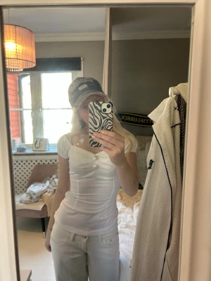 Vit topp med pärlknappar och puffärm - Söt vit topp med korta puffärmar och två stora pärlknappar framtill. Toppen har en v-ringad skärning och är figurnära med lätt glansigt material. Perfekt för dig som gillar feminina detaljer och en chic look.