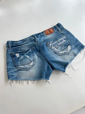 Blå jeansshorts från Fornarina - Säljer ett par blå jeansshorts från Fornarina med råa kanter och slitna detaljer. På ena bakfickan står det 'Fornarina hot chic' broderat i vitt. Klassisk femficksmodell med låg midja och knappgylf. Perfekta för sommaren!