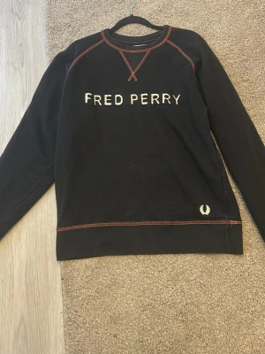 Svart sweatshirt från Fred Perry - Snygg svart sweatshirt från Fred Perry med kontrastsömmar i rött och broderad logga på bröstet samt det klassiska Fred Perry-emblemet nedtill. Tröjan har rund hals och långa ärmar. Perfekt för en casual och stilren look.