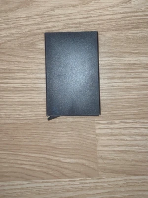 Svart minimalistisk korthållare - Snygg och stilren svart korthållare i metall med slimmad design. Perfekt för dig som vill ha ett enkelt och modernt sätt att förvara dina kort. Passar lätt i fickan och har en clean look utan synliga loggor eller detaljer.