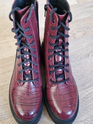 Vinröda kängor med krokopräglat mönster - Säljer ett par vinröda kängor med en krokodilskinn look och svarta snören. Kängorna har grov sula, dragkedja på sidan och är tillverkade i glansigt konstläder. Kommer ej ihåg vart dom orginellt är köpt ifrån.