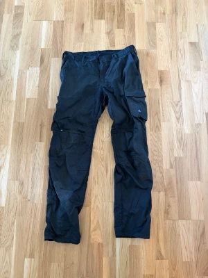 Svarta cargobyxor från Björnkläder - Snygga svarta cargobyxor från Björnkläder med flera praktiska fickor på benen och bak. Byxorna har en rak passform och är tillverkade i slitstarkt material, perfekta för dig som gillar en funktionell och stilren look. Nypris: 799kr 