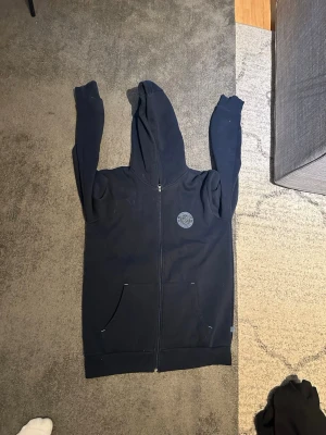 Mörkblå djurgårdshoodie med dragkedja - Säljer en mörkblå hoodie från Djurgården med dragkedja och huva. Den har två stora fickor framtill och ett diskret logomärke på bröstet. Perfekt för dig som gillar en enkel och stilren look. Materialet är mjukt och bekvämt.