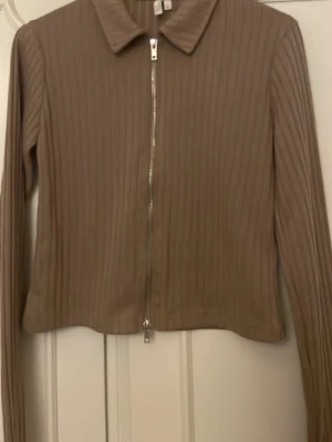 Beige ribbad ziptröja från NLY Trend - Säljer en beige ribbad tröja från NLY Trend med dragkedja framtill och klassisk krage. Tröjan har långa ärmar och är tillverkad i ett stretchigt material som sitter snyggt på kroppen. Perfekt att styla med jeans eller kjol.