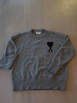 Grå stickad tröja från Ami - Snygg grå stickad tröja från Ami med svart hjärta och bokstaven A broderat på bröstet. Tröjan har rund halsringning, ribbade muddar och nederkant. Perfekt för dig som gillar stilrena och trendiga plagg. Väldigt fint skick och man känner verkligen kvalite i tröjan. Finns tags och qr kod där man direkt kommer in på amis hemsida. Storlek m men passar s också.