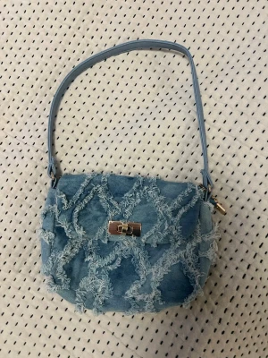 Ljusblå handväska i denim - Ljusblå handväska i denim med fransiga detaljer i rutmönster. Väskan har ett guldfärgat spänne framtill och en rem som går att ha över axeln. Supersöt, passar perfekt till jeans och läder, och är i perfekt skick. 