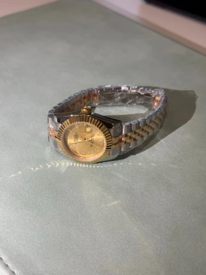 Rolx Datejust guld och silver klocka - Snygg Rolx Datejust med tvåfärgad länk i guld och silver. Klockan har en rund urtavla i guld med romerska siffror och räfflad bezel. Klassisk och tidlös design som lyfter vilken outfit som helst.