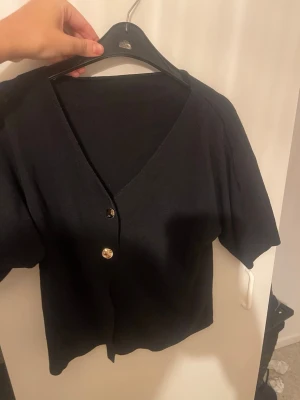 Svart kortärmad kofta med knappar - Svart kofta med korta ärmar och djup v-ringning. Två stora guldfärgade knappar framtill ger en snygg detalj. Tillverkad i ett mjukt, tunt material som känns skönt mot huden. Enkel och stilren modell som passar till många olika looks.