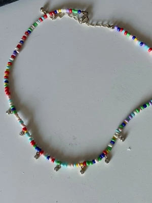 Färgglatt pärlhalsband med berlocker - Säljer ett halsband med små, runda pärlor i flera färger som rött, blått, grönt, vitt och gult. Halsbandet har små ” diamant ” berlocker som hänger längs kedjan. Perfekt för dig som gillar färgglada accessoarer och vill sticka ut lite extra.