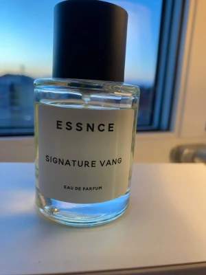 ESSNCE Signature Vang Eau de Parfum - Minimalistisk parfymflaska från ESSNCE med doften Signature Vang. Flaskan är cylindrisk i genomskinligt glas med svart rund kork och vit etikett med svart text. Innehållet är ljusgult och flaskan har en modern, stilren design. Mängd i flaskan enl bilder