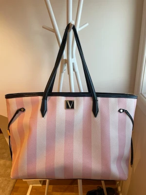 VS väska - Säljer en väska från Victoria Secret med breda rosa och vita ränder. Väskan har svarta handtag och detaljer samt ett metallmärke med V-logga framtill. Perfekt för dig som vill ha plats för allt och gillar en trendig look.
