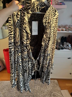 Zebra-mönstrad omlottklänning Zara - Zebra-mönstrad omlottklänning från Zara med långa ärmar och knyt i midjan. Klänningen har en djup v-ringning och volangdetaljer längs kanterna. Materialet är mjukt och följsamt, troligtvis polyester. Perfekt för dig som vill sticka ut med ett djuriskt mönster.