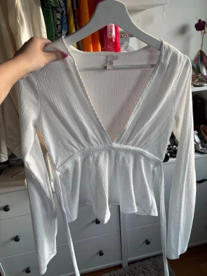 Vit omlottopp från Nelly - Trendig vit omlottopp från Nelly med ribbad struktur och långa ärmar. Toppen har en djup v-ringning och knytband i midjan för en snygg passform. Perfekt att styla med jeans eller kjol för en fräsch look.