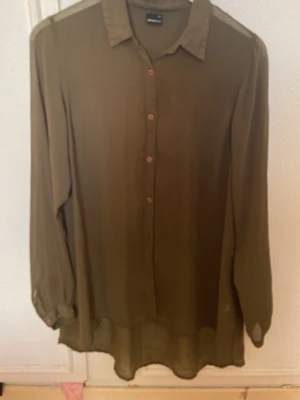 Olivgrön skjorta från Gina Tricot - Stilren olivgrön skjorta från Gina Tricot med klassisk krage och knappar framtill. Skjortan har lång ärm och en loose passform som ger en avslappnad vibe. Perfekt att styla lager på lager eller bära som den är.