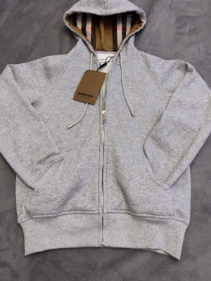 Grå zip hoodie från Burberry - Snygg grå zip hoodie från Burberry med klassiskt rutigt foder i huvan och dragsko. Tillverkad i mjuk bomullsblandning, har fickor fram och ribbade muddar. Perfekt för en avslappnad och stilren look.