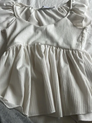 Vit  topp från 157 - Supersöt vit topp från 157 med volangärmar och ribbad struktur. Toppdn har en markerad midja och ett flowy fall som ger en romantisk vibe. Perfekt för dig som gillar en feminin och trendig stil.