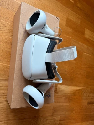 Oculus Quest 2 VR-headset med kontroller - Säljer ett Oculus Quest 2 VR-headset i vit plast med matchande vita kontroller. Headsetet har justerbart tygband och mjuk vaddering runt ögonen för extra komfort. Perfekt för gaming och VR-upplevelser hemma. Lite färg och boxen är sönder men annars inga problem funkar helt perfekt. Box medföljer!😀