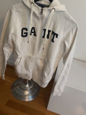 Vit hoodie med dragkedja från Gant - Vit hoodie från Gant med dragkedja framtill och stora blåa  bokstäver på bröstet. Klassisk huva med snören och två fickor framtill. Tillverkad i mjukt bomullsmaterial, perfekt för en avslappnad och sportig stil. En fläck baktill men kan gå bort!
