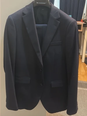 Mörkblå kostym med kavaj och byxor - Snygg mörkblå kostym med klassisk kavaj och matchande byxor. Kavajen har två knappar, smala slag och flera fickor framtill. Perfekt för dig som vill ha en stilren och tidlös look. Materialet känns mjukt och följsamt. Storlek är Slim fit. Byxor 48