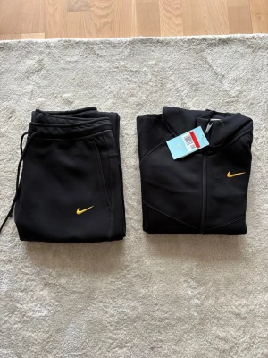 Svart Nike hoodie med gul logga - Snygg svart hoodie från Nike med dragkedja framtill och huva. Hoodien har en gul Nike-logga på bröstet och två fickor med dragkedja. Tillverkad i ett mjukt och stretchigt material som känns sportigt och modernt. Perfekt för dig som gillar streetwear och sportig stil.