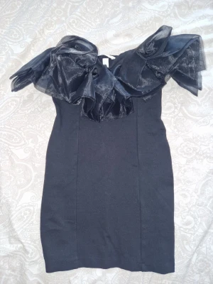 Svart kort klänning med volang från HM - Svart klänning med bandeau-design. Klänningen är figurnära och kort, perfekt för dig som vill ha en elegant och unik look. Materialet är troligtvis polyester och volangerna har en snygg glansig finish. Super cute & använd en gång på bröllop💥😍