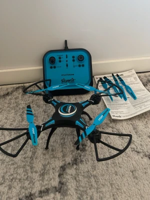 Blå och svart drönare Silverlit - Cool drönare från Silverlit i svart och blått med matchande fjärrkontroll. Kommer med extra propellrar och manual. Drönaren har skydd runt propellrarna och är perfekt för att flyga både inomhus och utomhus.knappt används 