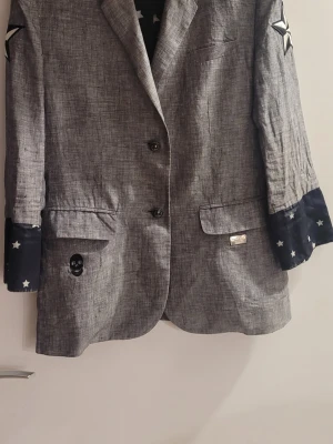 Grå Philipp Plein kavaj med Mickey Mouse - Unik grå kavaj från Philipp Plein med coolt Mickey Mouse-motiv och texten 'Bad Boy' på ryggen. Svarta detaljer med stjärnor på ärmsluten, dödskalle på framsidan och stjärnor på axlarna. Perfekt för dig som vill sticka ut med en edgy och lekfull stil.