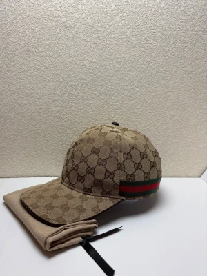 Beige keps med GG-mönster - Snygg beige keps med klassiskt GG-monogram över hela kepsen. På sidan finns den ikoniska gröna och röda randen. Kepsen har böjd skärm och justerbar rem bak. Tillverkad i canvasmaterial som ger en exklusiv känsla.