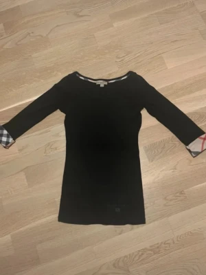 Svart tröja med rutiga detaljer - Svart tröja från Burberry med trekvartsärm och klassiska rutiga detaljer på ärmsluten. Tröjan har en enkel, rak passform och rund halsringning. Perfekt för dig som gillar stilrena plagg med en twist.