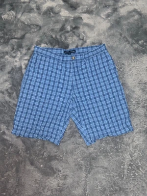 Tommy Hilfiger Rutiga Chino-shorts (Nyskick) -  ett par klassiska rutiga shorts från Tommy Hilfiger i olika blå nyanser. Sjukt snygga till sommarn! Oanvända och nytvättade med perfekt passform. • Material: Bomull  • Skick: 10/10 – Nyskick, inga defekter. • Detaljer: Gylf med knapp och dragkedja, fickor både fram och bak. 