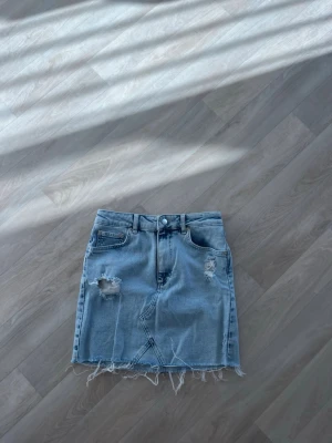 Ljusblå jeanskjol från NA-KD - Trendig ljusblå jeanskjol från NA-KD med slitna detaljer och fransig nederkant. Kjolen har klassiska fem fickor, knapp och dragkedja framtill. Perfekt för dig som vill ha en cool och avslappnad stil.