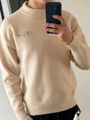 Beige stickad tröja Âme Dorée - Snygg beige stickad tröja från Âme Dorée med broderad logga på bröstet och stor logga i grått på ryggen. Tröjan har ribbade muddar, hög krage och är tillverkad i mjukt material. Skick 9/10, då jag ska försöka ta bort den noppiga innan den sälj, men det är med materialet. Priset är ej hugget i sten, mvh Erik