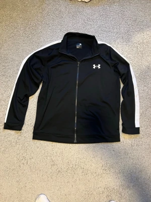 Svart träningsjacka från Under Armour - Snygg svart träningsjacka från Under Armour med hel dragkedja och vita ränder längs ärmarna. Jackan har ståkrage och det klassiska UA-logotypen på bröstet. Tillverkad i ett mjukt och stretchigt funktionsmaterial som passar perfekt till träning eller uppvärmning.