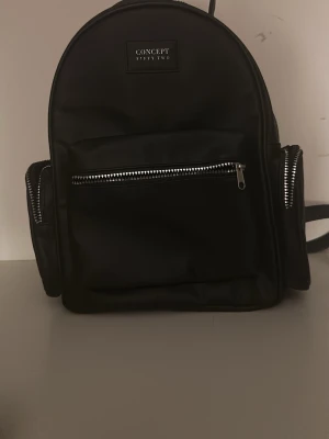 Svart ryggsäck Concept Fiftytwo - Snygg svart ryggsäck från Concept Fiftytwo med flera fack och silverfärgade dragkedjor. Tillverkad i ett glansigt material som ger en modern look. Perfekt storlek för att bära det viktigaste och har justerbara axelband för bästa komfort.