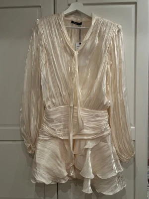 Fest klänning  - Superfin kort klänning i ljus, skimrande siden med detaljer och volang kjol. Klänningen har långärmade ärmar, knytband i halsen och markerad midja. Helt oanvänd med lappen kvar, nypris 600!!! Perfekt för dig som vill sticka ut med en elegant och trendig look. Hör av dig vid frågor. 