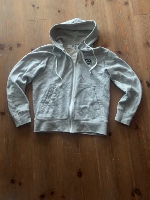 Grå hoodie Comme des Garçons Play - Grå hoodie från Comme des Garçons Play med dragkedja, huva och vita snören. Klassisk svart hjärtlapp med ögon på bröstet. Tillverkad i mjukt bomullsmaterial och har två fickor framtill. Perfekt för en avslappnad och stilren look.