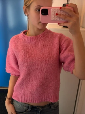 Rosa stickad tröja med kort ärm - Säljer en söt rosa stickad tröja med korta ärmar och ribbade muddar vid hals, ärmslut och nederkant. Tröjan har en avslappnad passform och är gjord i ett mjukt, fluffigt material som känns skönt mot huden. Perfekt för dig som gillar färg och vill ha en mysig look.
