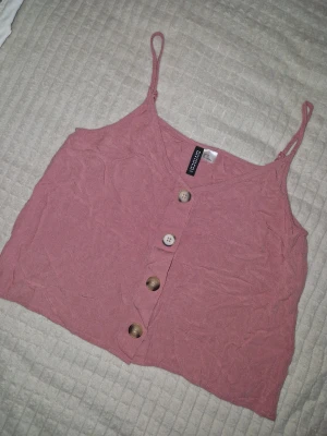 Rosa linnetopp från H&M Divided - Söt rosa linnetopp från H&M Divided med smala axelband och dekorativa knappar framtill. Toppen har en croppad passform och är tillverkad i ett mjukt bomullsmaterial. Legat nerpackad därav skrynklig 🌞