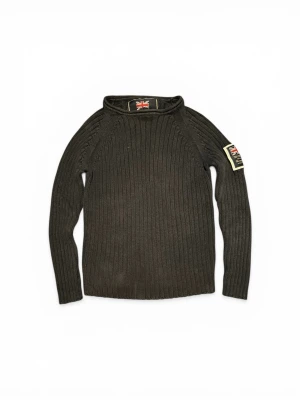 Mörkgrön ribbstickad tröja Morris - Mörkgrön ribbstickad tröja från Morris med lång ärm och patch med brittisk flagga på ena ärmen. Tröjan har en högre krage och en klassisk, lite militärinspirerad look. Perfekt för dig som gillar stilrena och tidlösa plagg.