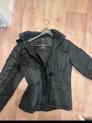 Svart regnkappa med huva - Snygg svart regnjacka med huva och dragkedja framtill. Jackan har flera fickor, både med lock och dragkedja, samt en figurnära passform. Perfekt för kalla dagar och enkel att matcha med olika outfits. Materialet är glansigt och quiltat för extra värme.