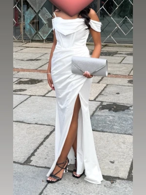 Vit axelbandslös balklänning med slits - Elegant vit långklänning med offshoulder och smickrande drapering upptill. Klänningen har en hög slits framtill och figurnära passform. Perfekt för bal eller student. Materialet har en lätt glansig finish som ger en lyxig känsla. Köpt på dressroomlinne, ordinare pris 3980kr